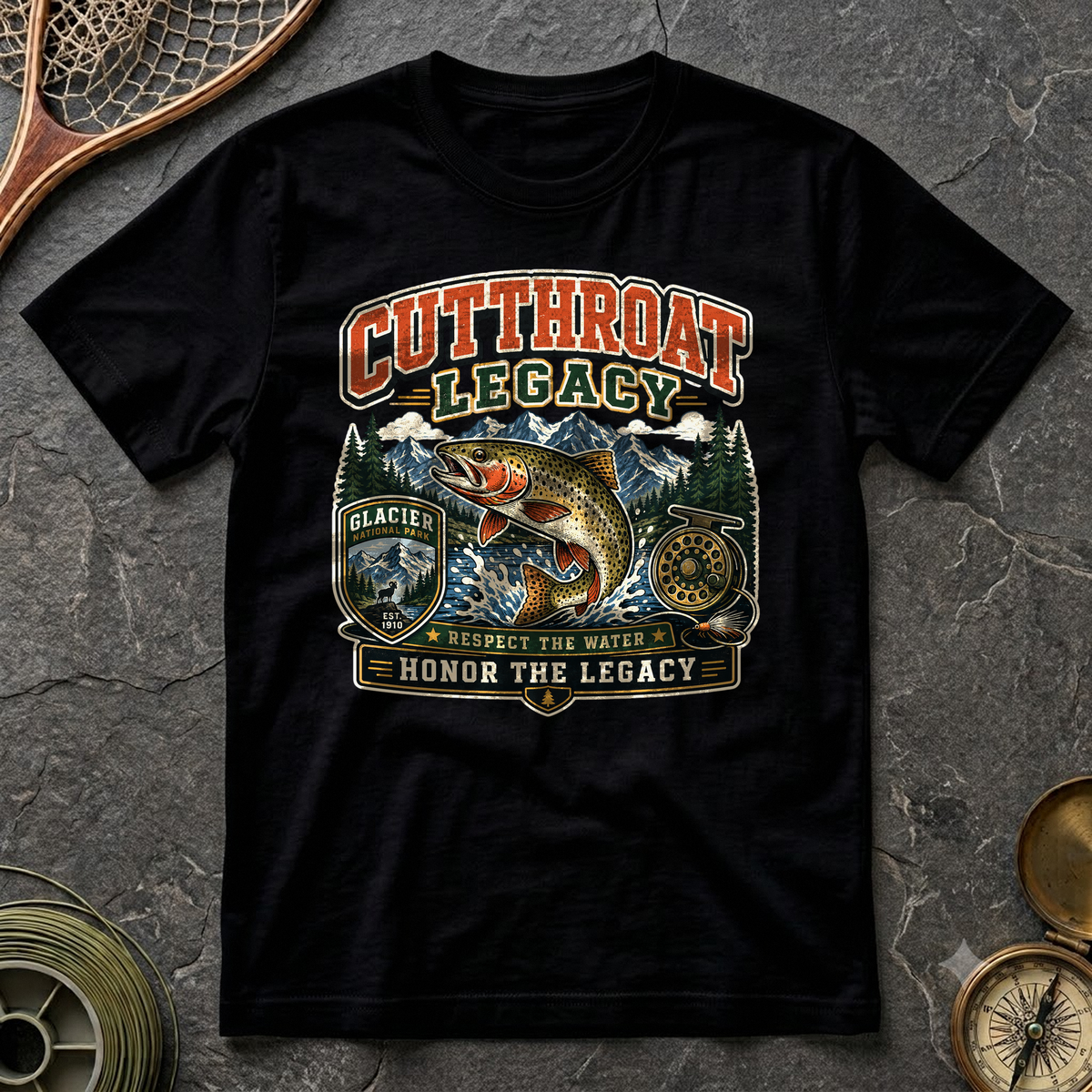 Cutthroat Legacy tee in black, flat-lay on dark slate with a wooden landing net, brass compass and a spool of tippet