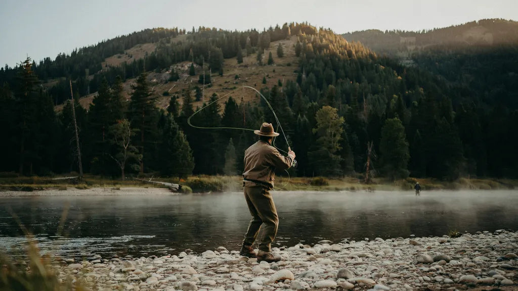 An angler mid-backcast on a misty mountain river at sunrise, the fly line unrolling in a loop above and behind the rod tip, with a pine ridge and pale gravel bar in first light