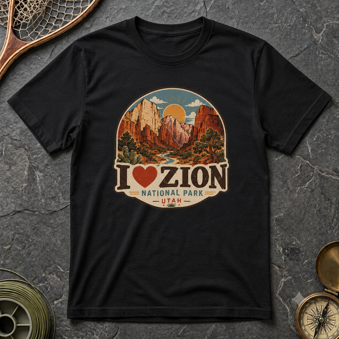 A black short-sleeve graphic T-shirt laid flat on a dark stone surface, featuring a circular scenic print of Zion National Park with the text 'I ♥ ZION NATIONAL PARK - UTAH.' Decorative props including a fishing net, a spool, and a compass are partially visible around the shirt.
