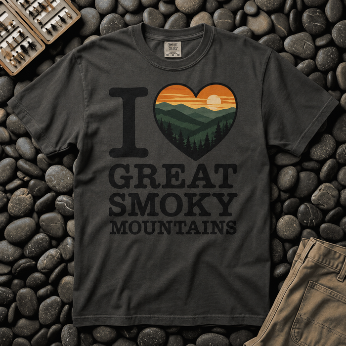 A dark charcoal gray t-shirt laid flat on a bed of dark river stones, featuring a graphic that reads 'I [heart with a sunset mountain landscape] GREAT SMOKY MOUNTAINS'.