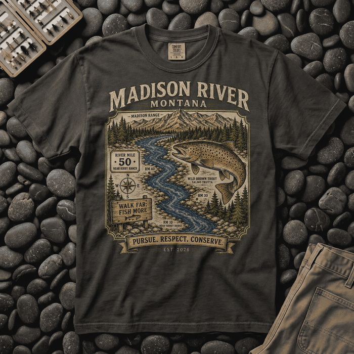 Pepper Comfort Colors Madison River Montana trout t-shirt laid flat on river stones beside a fly box, showing the river map and leaping brown trout design