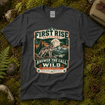 Dark heather grey Answer the Call of the Wild tee laid flat on green moss with ferns, river stones and a pinecone, showing a vintage badge of a howling wolf under a full moon — thumbnail 1