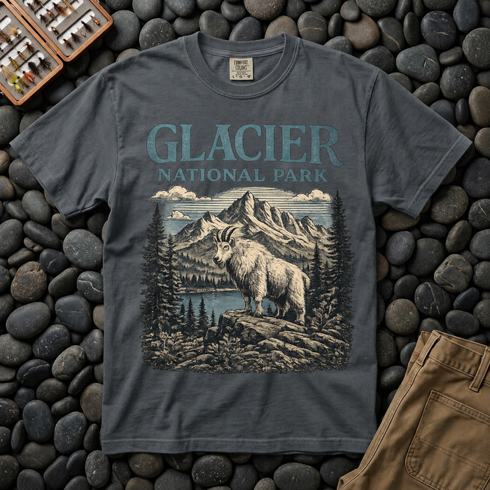 Dark gray Glacier National Park mountain goat t-shirt laid flat on river stones beside a fly box and canvas pants