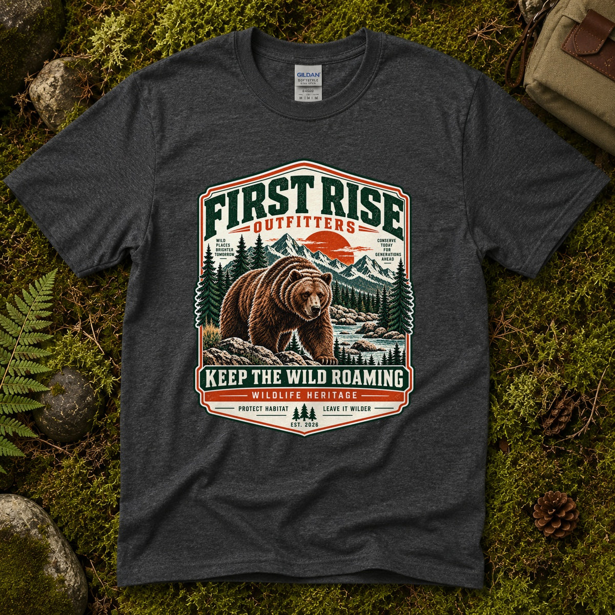 Dark heather grey Keep the Wild Roaming tee laid flat on green moss with ferns, river stones and a pinecone, showing a vintage badge of a grizzly bear above a mountain river and pines