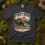 Dark heather grey Keep the Wild Roaming tee laid flat on green moss with ferns, river stones and a pinecone, showing a vintage badge of a grizzly bear above a mountain river and pines — thumbnail 1