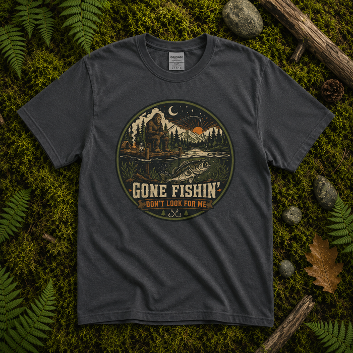 A dark charcoal gray t-shirt laid flat on a bed of green moss, ferns, and small rocks. The t-shirt features a circular graphic print in the center with a night outdoor scene showing a person fishing by a lake, a moon, and trees, with the text 'GONE FISHIN' I DON'T LOOK FOR ME'.