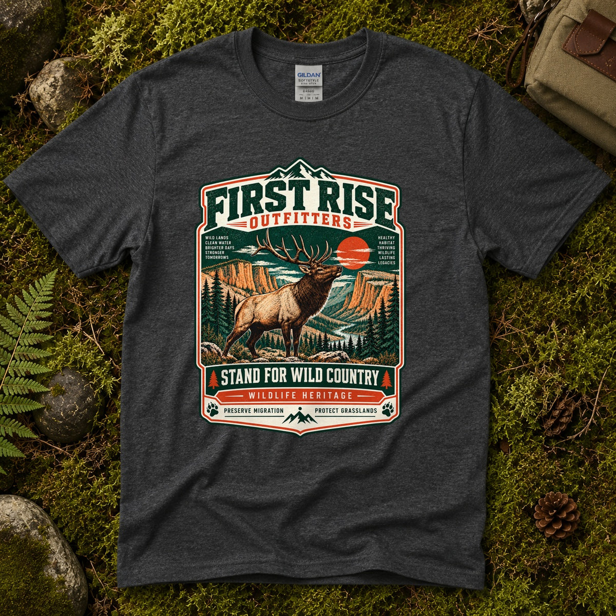 Dark heather grey Stand For Wild Country tee laid flat on green moss with ferns, river stones and a pinecone, showing a vintage badge of a bugling bull elk above canyon country and pines