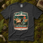 Dark heather grey Stand For Wild Country tee laid flat on green moss with ferns, river stones and a pinecone, showing a vintage badge of a bugling bull elk above canyon country and pines — thumbnail 1